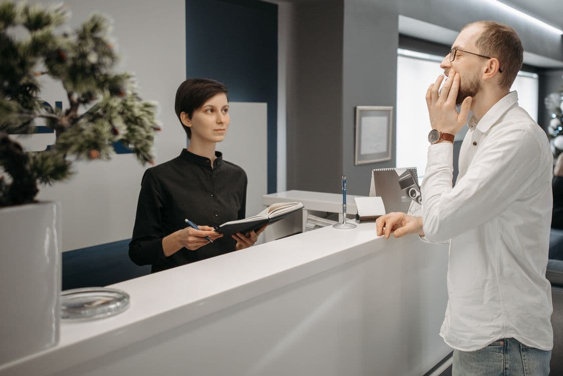 Patient holding his jaw in pain while booking an urgent dental appointment at the reception desk